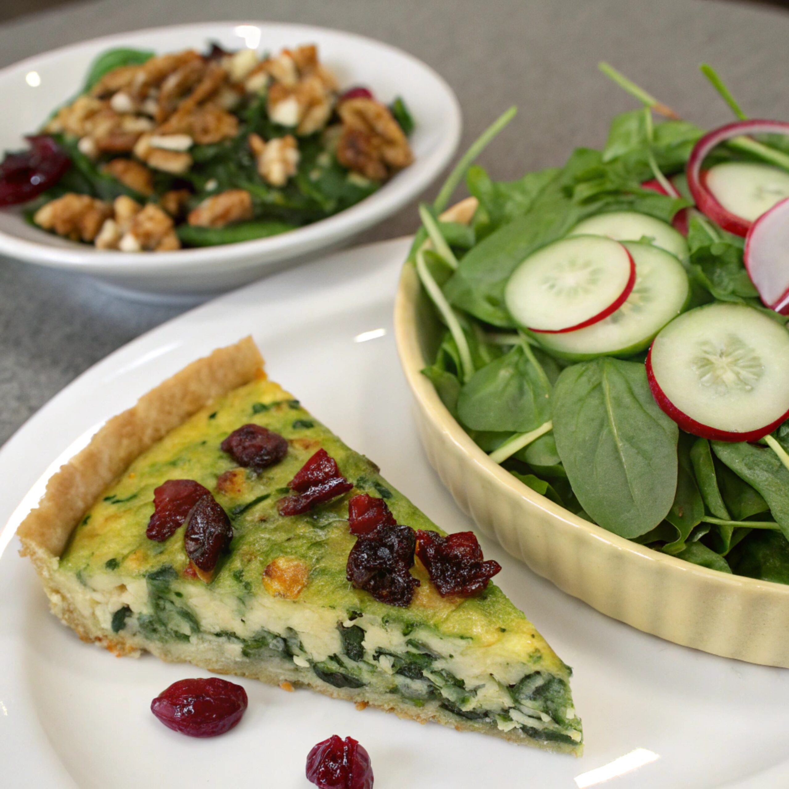 A serving of spinach quiche plated alongside a vibrant green salad with mixed greens, arugula, and baby kale, garnished with sliced radishes, cucumbers, nuts, and dried cranberries, drizzled with vinaigrette.