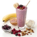 A tall glass filled with a purple berry smoothie, topped with a banana slice and mixed berries. Surrounding the glass are ingredients including a banana, almond butter, Greek yogurt, mixed nuts, and frozen berries.