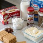 Ingredients for no-bake cheesecake including cream cheese, graham crackers, whipped cream, and flavorings laid out