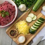 Raw ingredients for Ground Beef Zucchini Bake including minced beef, sliced zucchini, shredded cheese, chopped onions, garlic, and herbs.