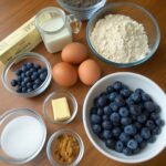 An arrangement of ingredients including fresh blueberries, flour, sugar, butter, eggs, vanilla extract, and spices for making Blueberry Crunch Muffins.