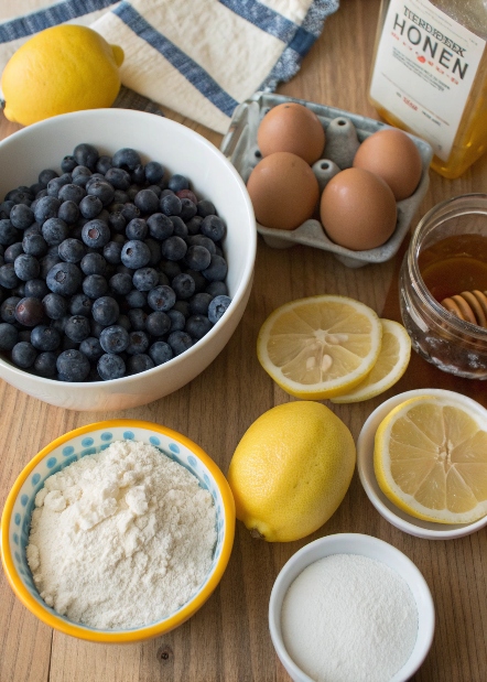 Assorted fresh ingredients for gluten-free blueberry lemon cake, including blueberries, lemons, gluten-free flour, eggs, and honey