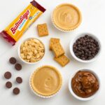 Assorted ingredients for Butterfinger Caramel Crunch Mini Pies, including Butterfinger bars, pie crusts, caramel sauce, chocolate chips, and peanut butter.